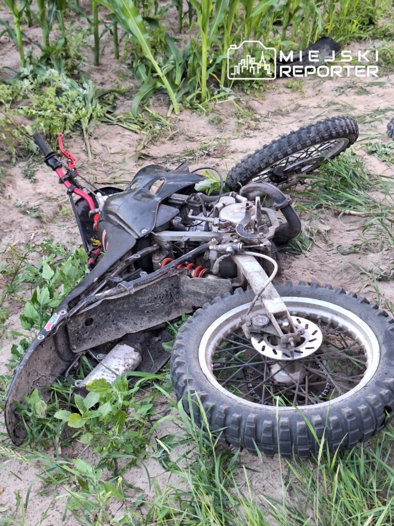 Motocykl leży na boku w zaroślach, otoczony wysoką trawą i roślinami.