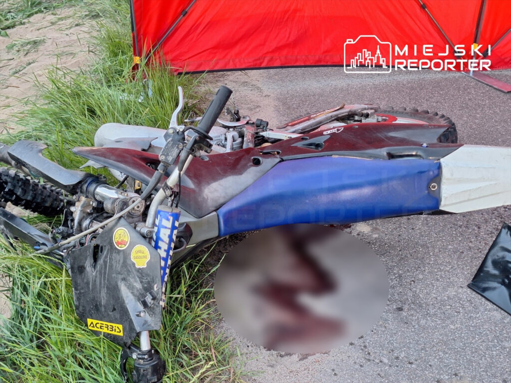 Motocykl terenowy leży na ziemi w trawie, obok czerwonego namiotu, z widocznymi uszkodzeniami.