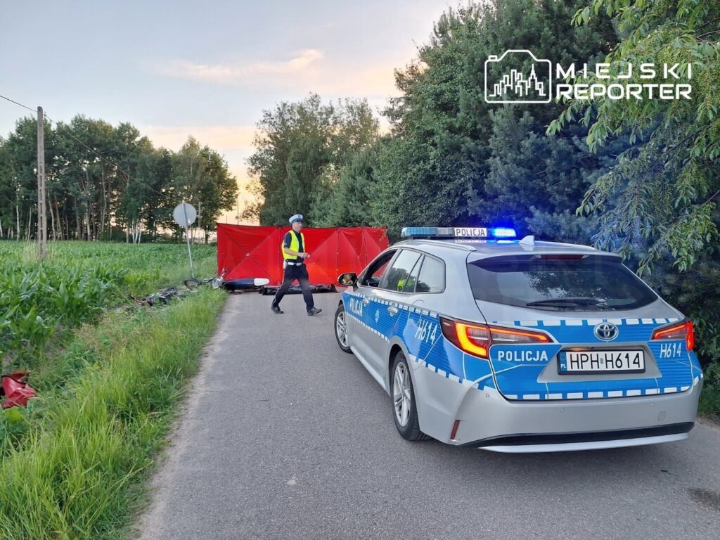 Policjant w kamizelce odblaskowej przechodzi obok policyjnego samochodu przy zamkniętej drodze, otoczonej drzewami.