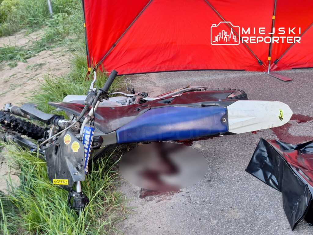 Motocykl terenowy leży na poboczu drogi, obok widoczne plamy krwi oraz czarna folia. W tle czerwony namiot.