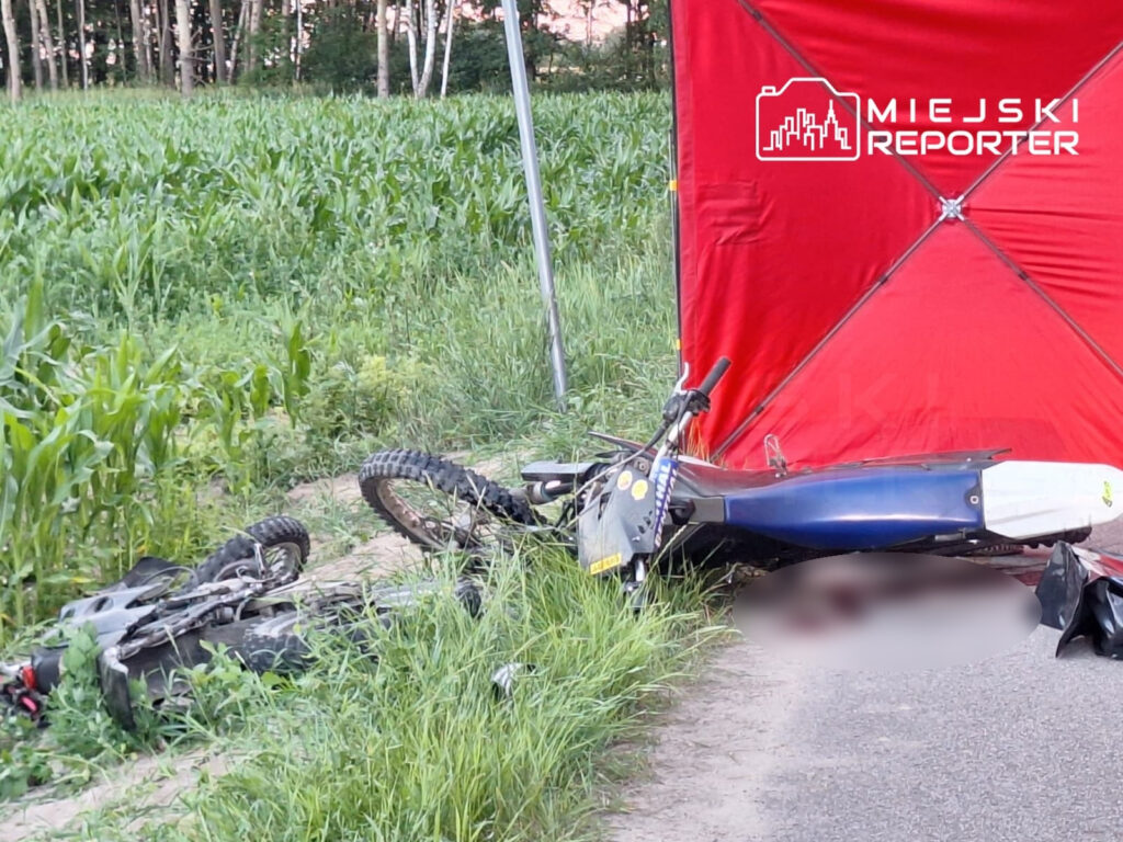 Dwa motocykle leżą na poboczu drogi, otoczone wysoką trawą, z czerwonym namiotem w tle.