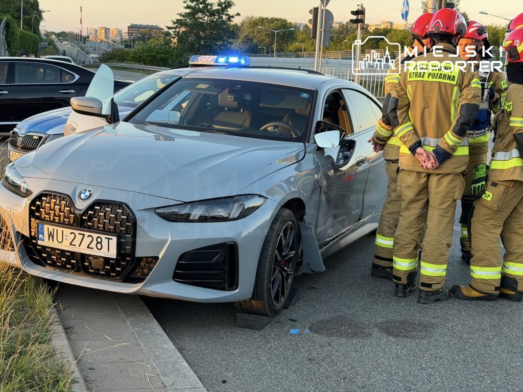 Grupa strażaków w mundurach omawia sytuację przy uszkodzonym samochodzie osobowym na poboczu drogi.