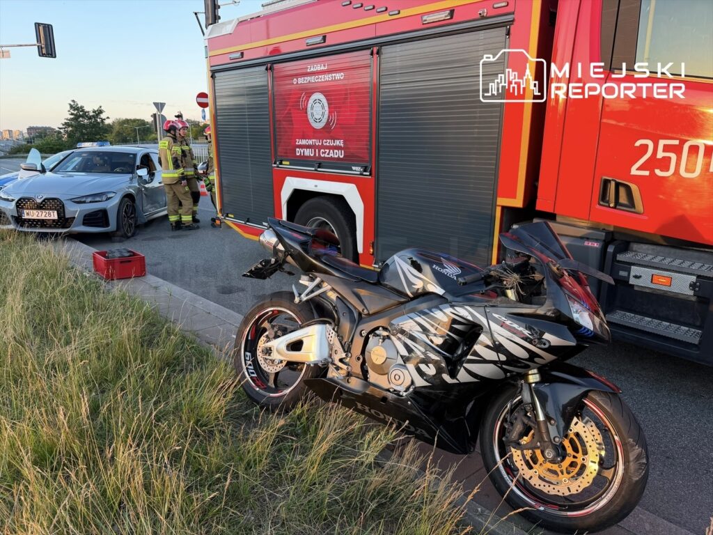 Na poboczu drogi stoi uszkodzony motocykl, obok którego pracują strażacy, a w tle widoczny jest samochód osobowy.