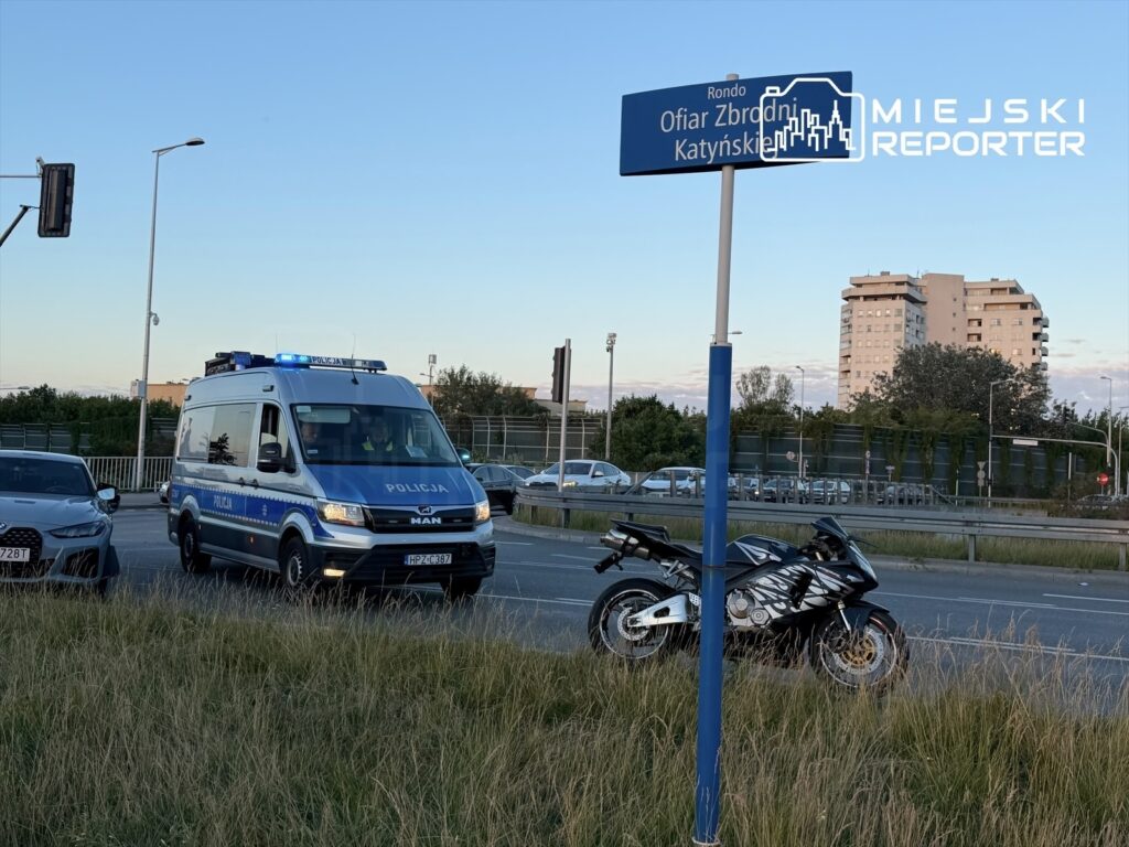 Na rondzie Ofiar Zbrodni Katyńskiej stoi policyjny radiowóz z włączonymi sygnałami świetlnymi obok motocykla.