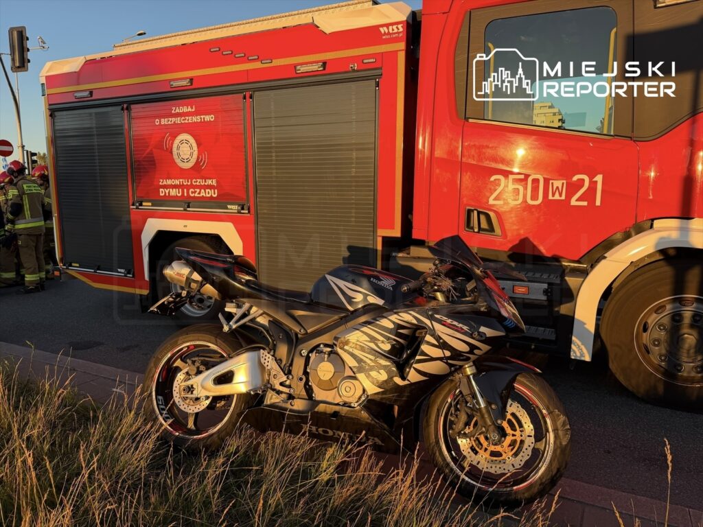 Motocykl sportowy stoi obok wozu strażackiego, a w tle widać strażaków przygotowujących się do akcji.