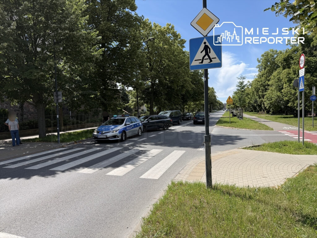 Policyjny radiowóz zatrzymany przy przejściu dla pieszych na ulicy otoczonej drzewami i parkingiem.