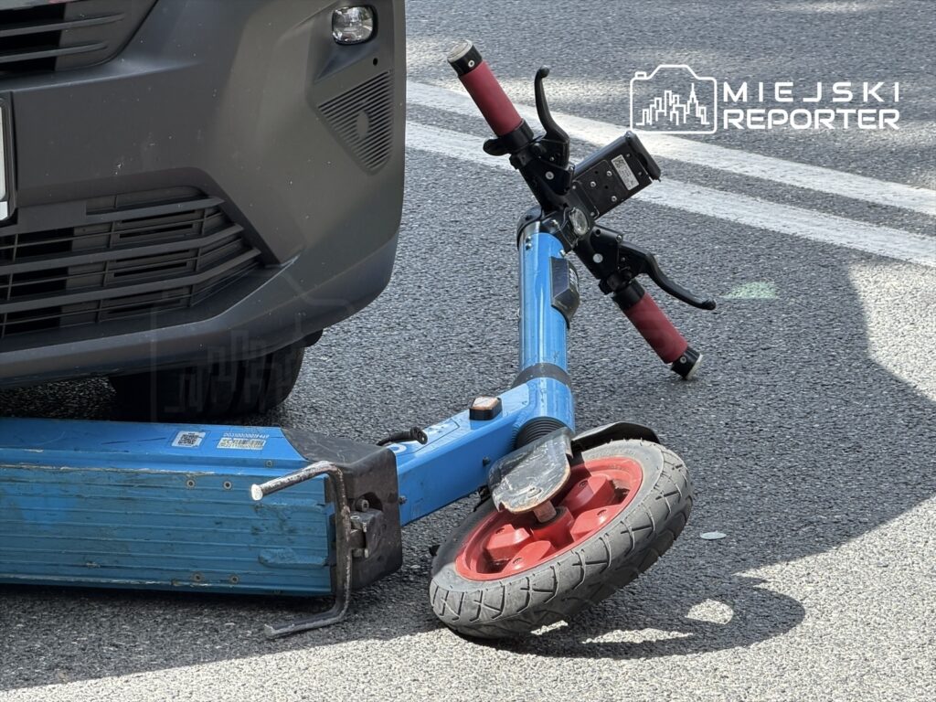 Złamana hulajnoga elektryczna leży na jezdni obok przedniego zderzaka samochodu.
