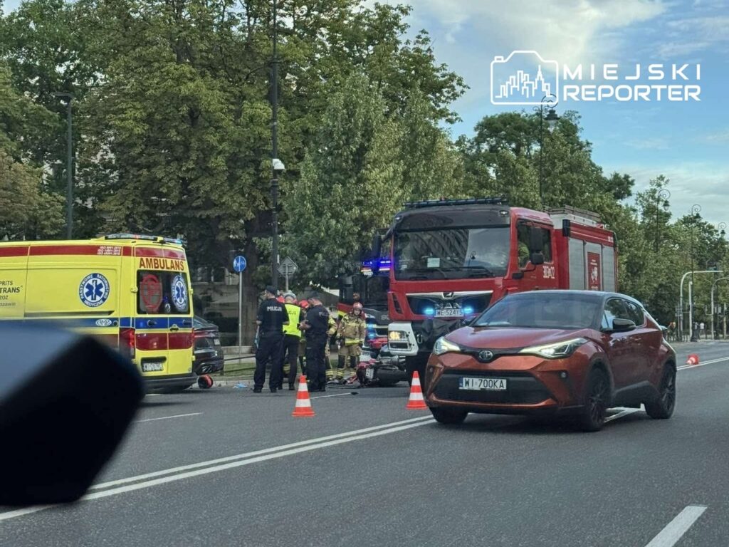 Zespół ratunkowy z ambulansu i straży pożarnej interweniuje na miejscu wypadku, otoczony przez policję.