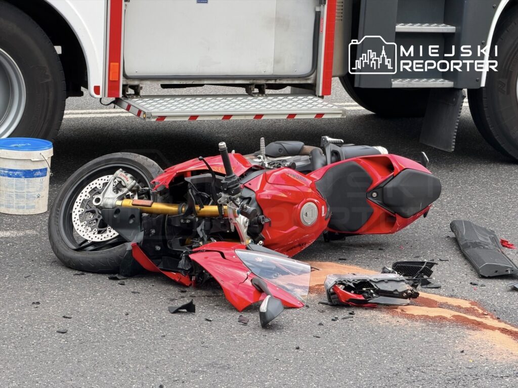 Czerwony motocykl leży na boku na jezdni, obok niego widoczne są rozbite elementy oraz plama oleju.