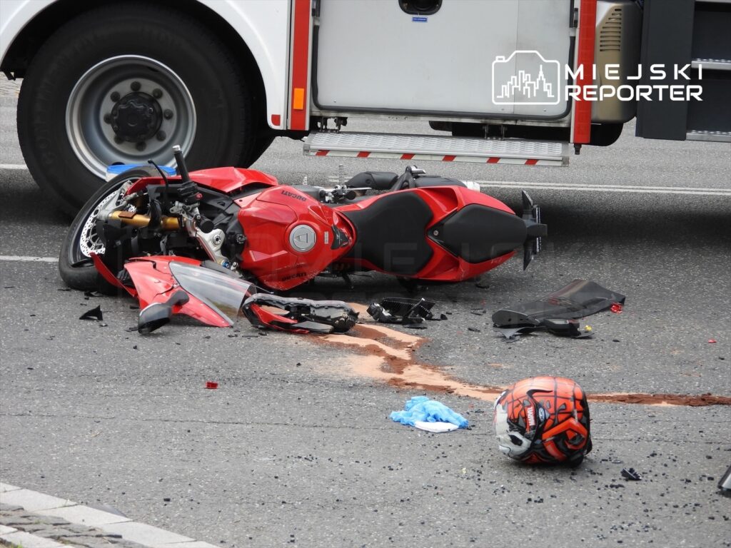 Zniszczony motocykl leży na jezdni, obok niego czerwona kask i plama płynu, w tle widać pojazd ratunkowy.
