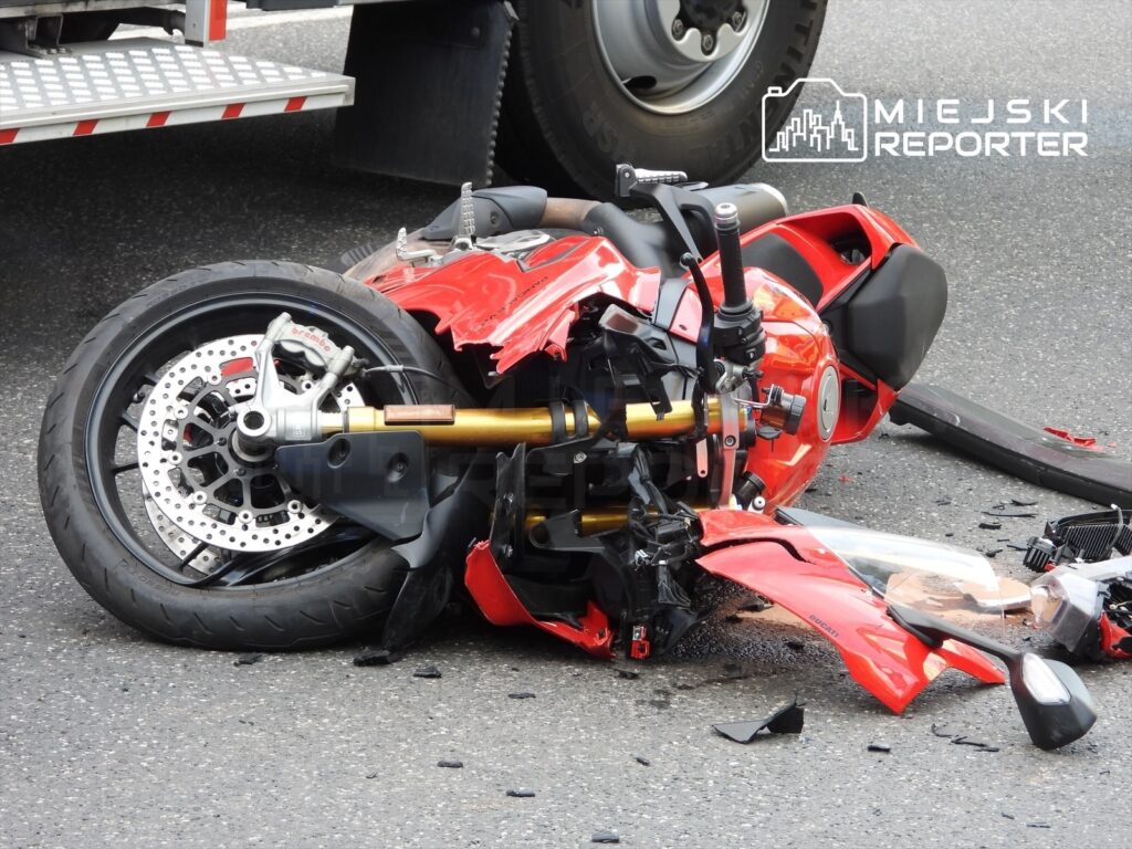 Uszkodzony motocykl leży na drodze, z rozbitym nadwoziem i odłamkami plastiku wokół.