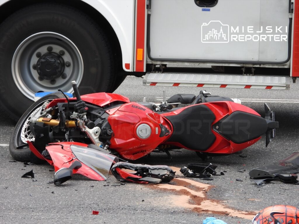 Czerwony motocykl leży na jezdni, obok widoczny jest duży pojazd ratunkowy. Na asfalcie widać uszkodzone elementy motocykla oraz plamę.