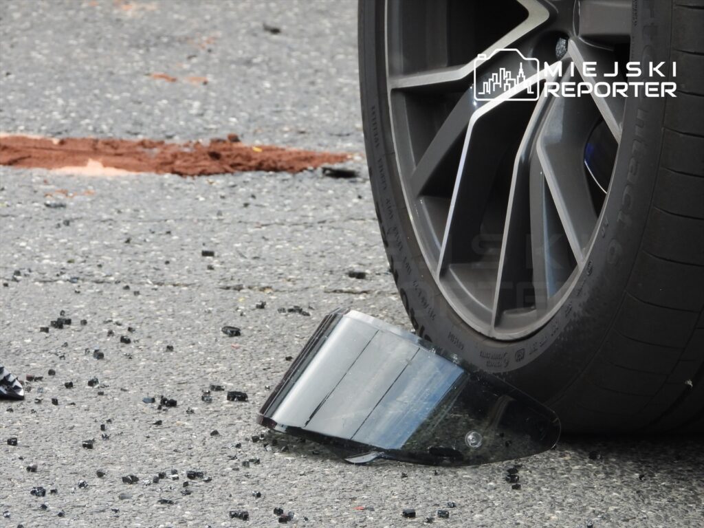 Złamany lusterko samochodowe leży na drodze obok opony pojazdu, a wokół rozrzucone fragmenty szkła.