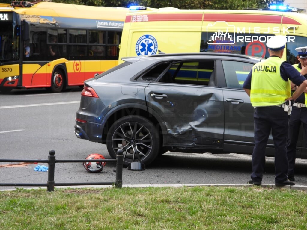 Czarny samochód osobowy z uszkodzeniami na boku stoi przy drodze, obok policjanci rozmawiający ze sobą.