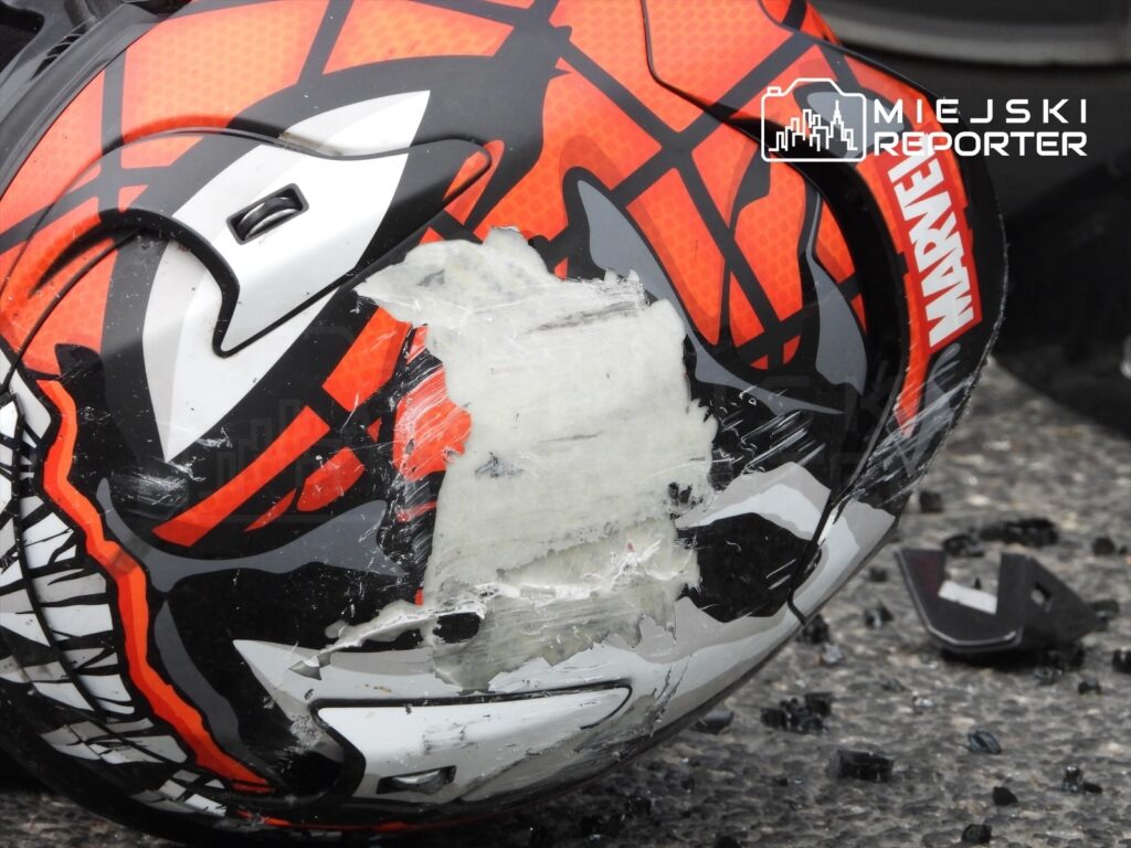 Zniszczony kask motocyklowy z motywem Spider-Mana leży na asfalcie, obok fragmenty szkła.