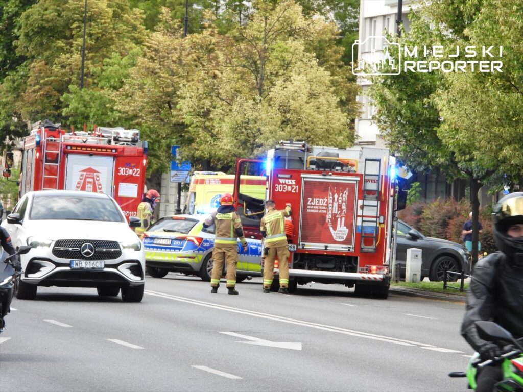 Ratownicy w hełmach i uniformach pracują przy wozie strażackim na ulicy, obok policyjnego radiowozu i samochodów osobowych.