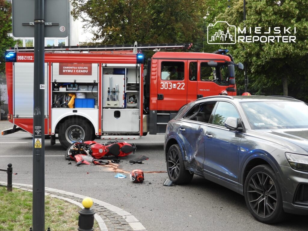 Czerwony wóz strażacki stoi obok miejsca wypadku, gdzie leży uszkodzony motocykl i kask. Obok stoi szary samochód z uszkodzoną karoserią.