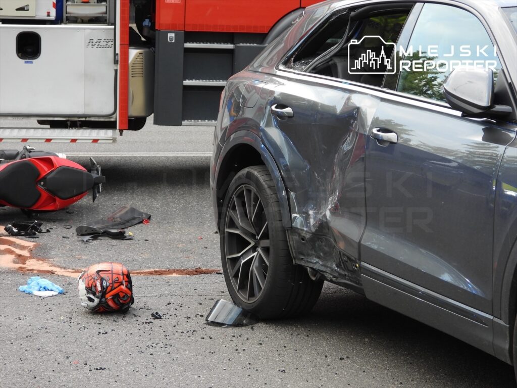 Motocykl leży na jezdni obok uszkodzonego samochodu osobowego, z rozbitym bokiem i plamą krwi na asfalcie.