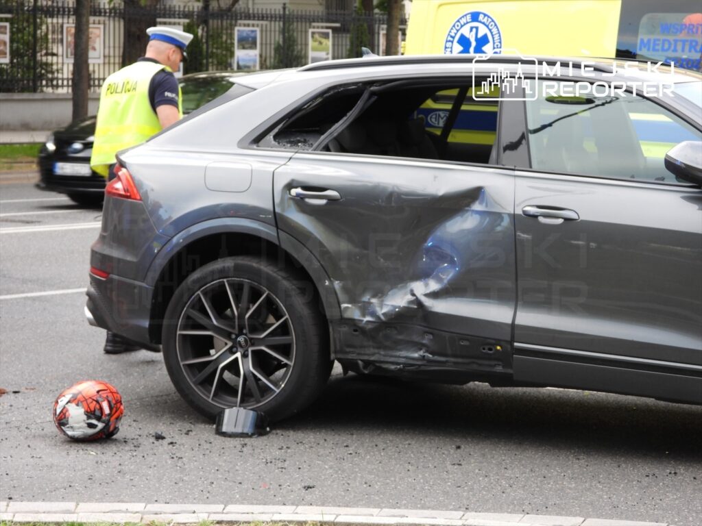 Policyjny funkcjonariusz sprawdzający uszkodzone auto z wgniecioną karoserią na ulicy, obok leży piłka.