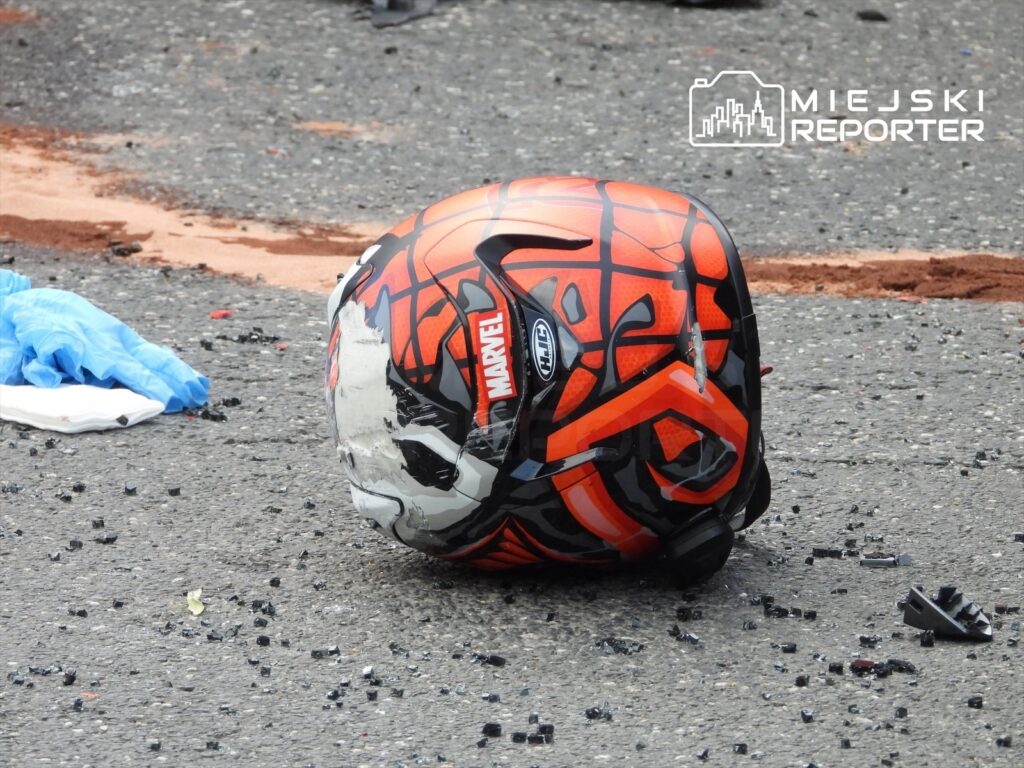 Kask motocyklowy w kolorze pomarańczowym z motywem Spider-Mana leży na asfalcie w otoczeniu rozbitego szkła.