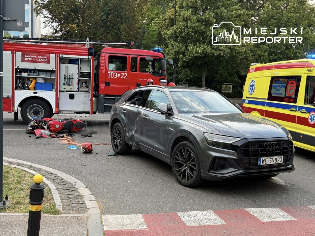 Motocykl leży na jezdni obok rozbitego samochodu, a w tle widoczne są wozy strażackie i ambulans.