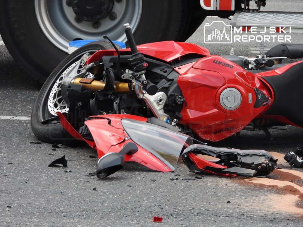 Czerwony motocykl leży na boku na drodze, obok niego rozrzucone fragmenty plastików i olej.