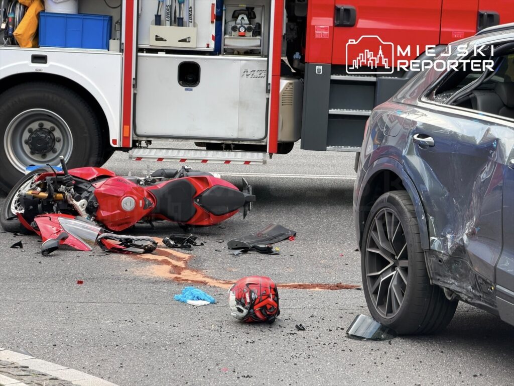 Motocykl leży na jezdni obok czerwonej karetki, a obok widoczne są rozbite elementy oraz plama krwi.