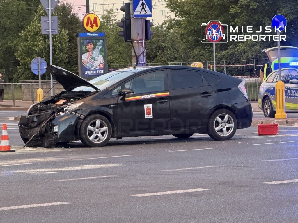 Czarny samochód osobowy z uszkodzoną przednią częścią stoi na skrzyżowaniu, obok oznakowanie drogowe i sygnalizacja.