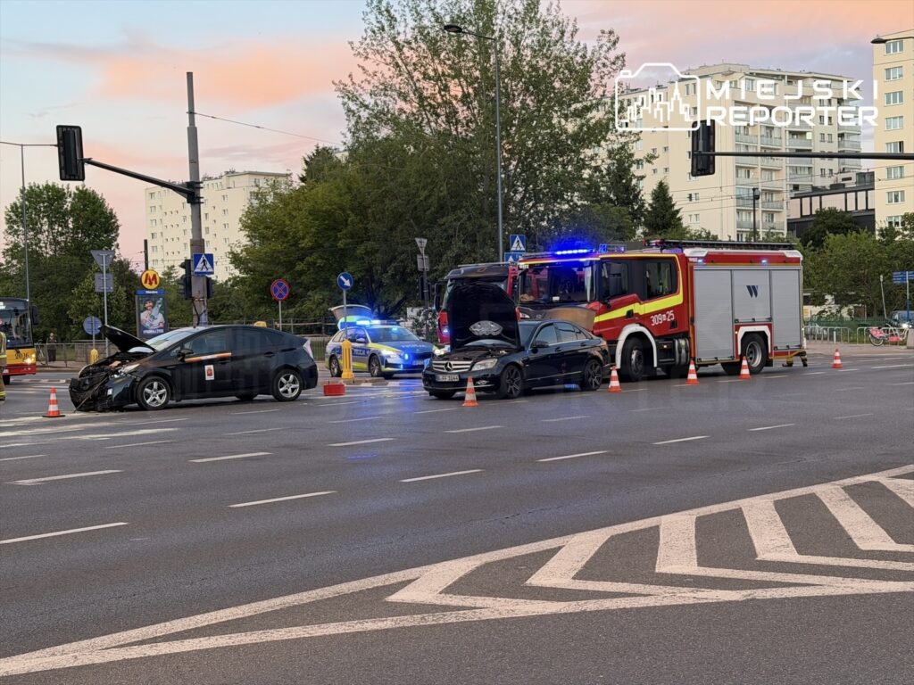 Czarny samochód osobowy z uszkodzoną maską stoi na ulicy, obok niego interweniuje straż pożarna oraz policja.