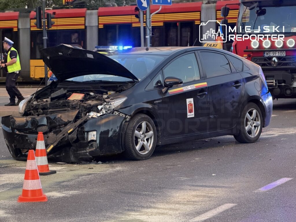 Uszkodzony taksówkarz z otwartą maską stoi obok pojazdu na ulicy, podczas gdy policjant kieruje ruchem.