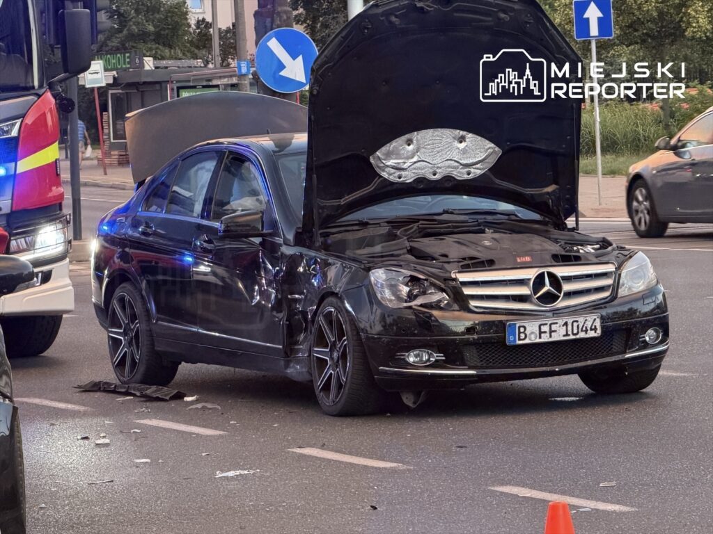 Czarny samochód osobowy z otwartą maską i uszkodzeniami na boku stoi na drodze, obok straż pożarna.