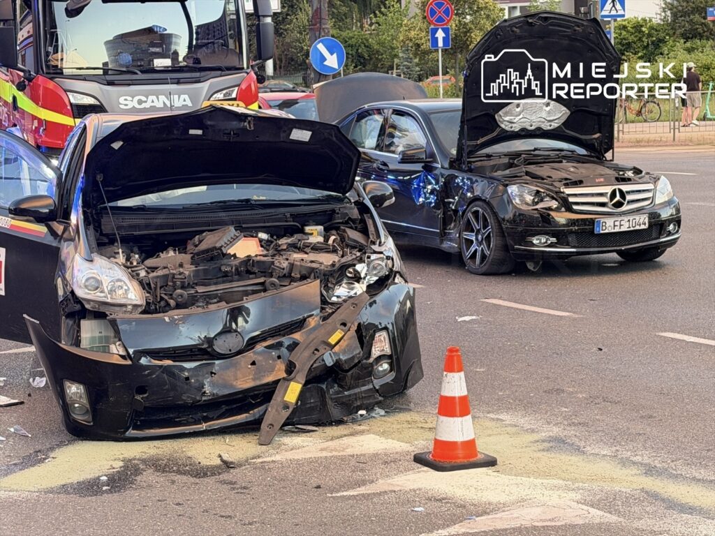 Dwa uszkodzone samochody osobowe na drodze, z widocznymi uszkodzeniami przodu, obok pomarańczowy słupek ostrzegawczy. W tle widać straż pożarną.