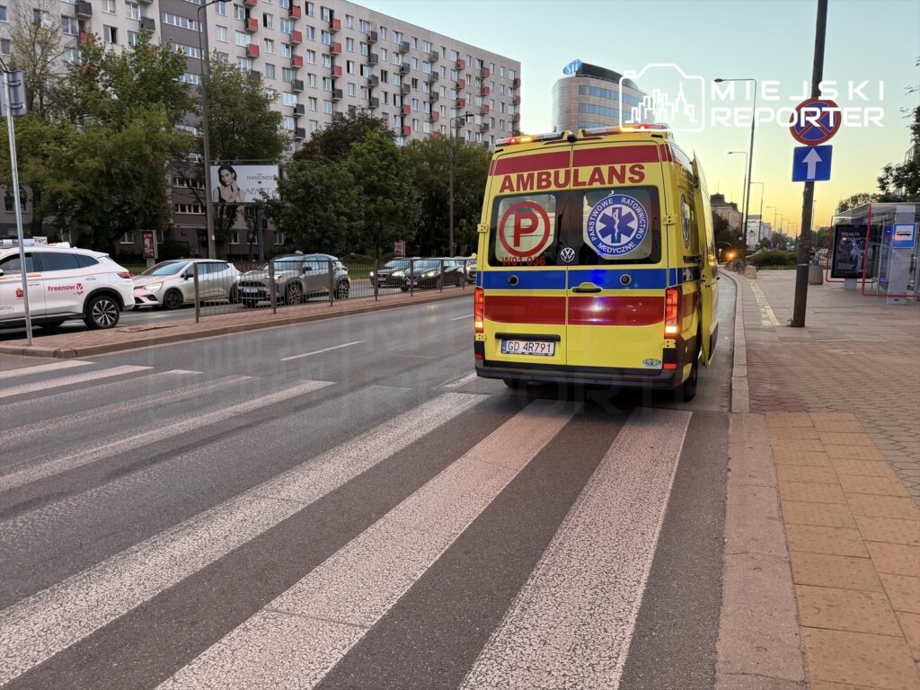 Karetka pogotowia ratunkowego zaparkowana na pasach w mieście, obok stojących samochodów i billboardu.