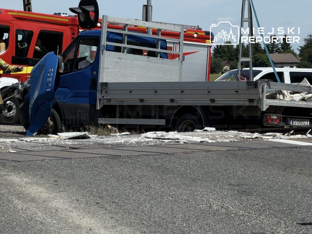 Niebieski samochód dostawczy z uszkodzoną kabiną stoi obok wozu strażackiego na drodze, wokół leżą fragmenty materiałów budowlanych.