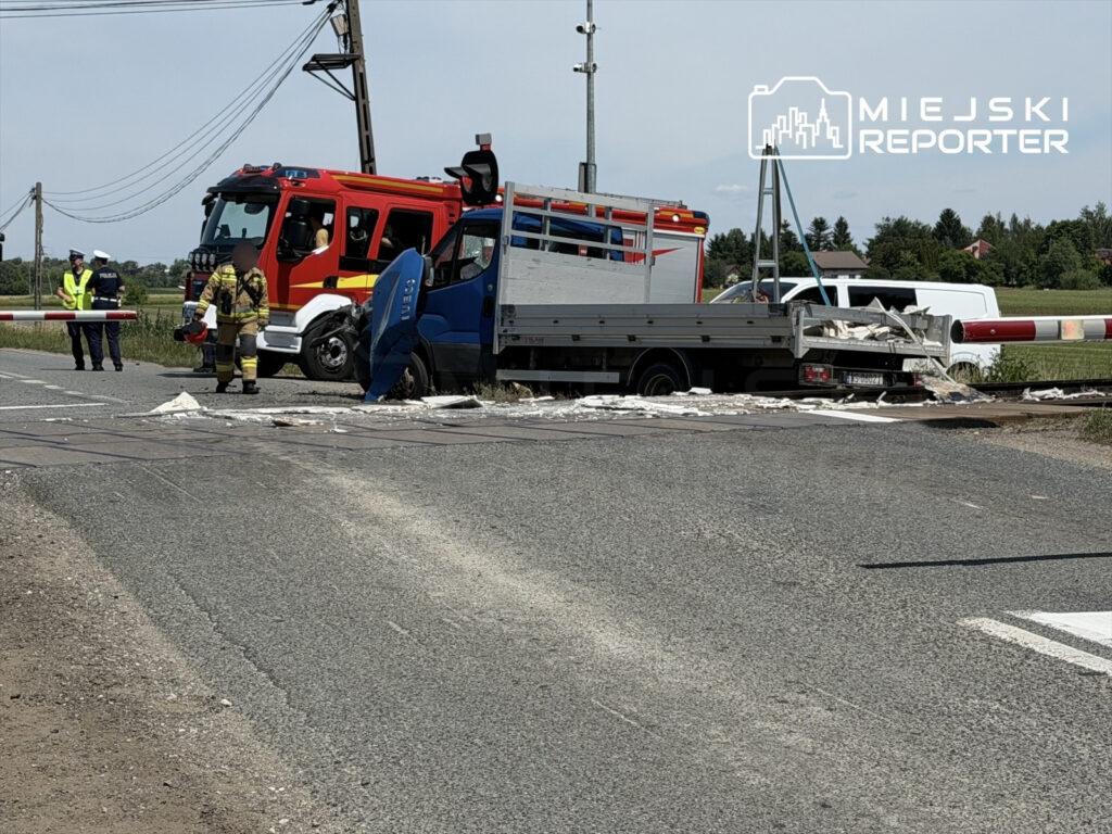 Strażacy i policjanci pracują przy wypadku drogowym obok torów kolejowych, gdzie stoi uszkodzony niebieski samochód dostawczy.