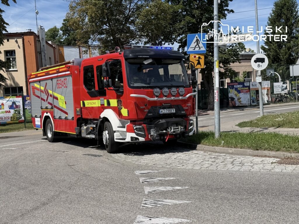 Czerwony wóz strażacki z włączonymi sygnalizatorami stoi na skrzyżowaniu, obok znaków drogowych i budynków.