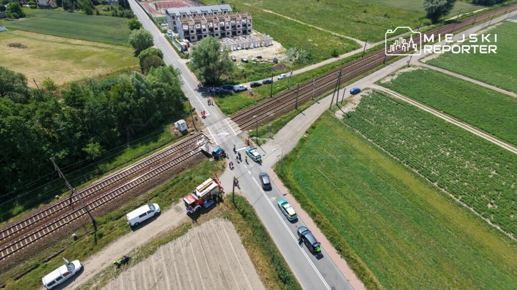 Ruch drogowy zablokowany na przejeździe kolejowym, w pobliżu nowo budowanych mieszkań. W tle widoczne pola i torowisko.