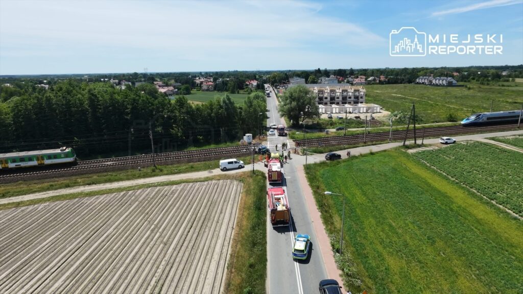 Na drodze w pobliżu przejazdu kolejowego zatrzymały się pojazdy ratunkowe, a w tle widać pociąg oraz budowę nowych domów.