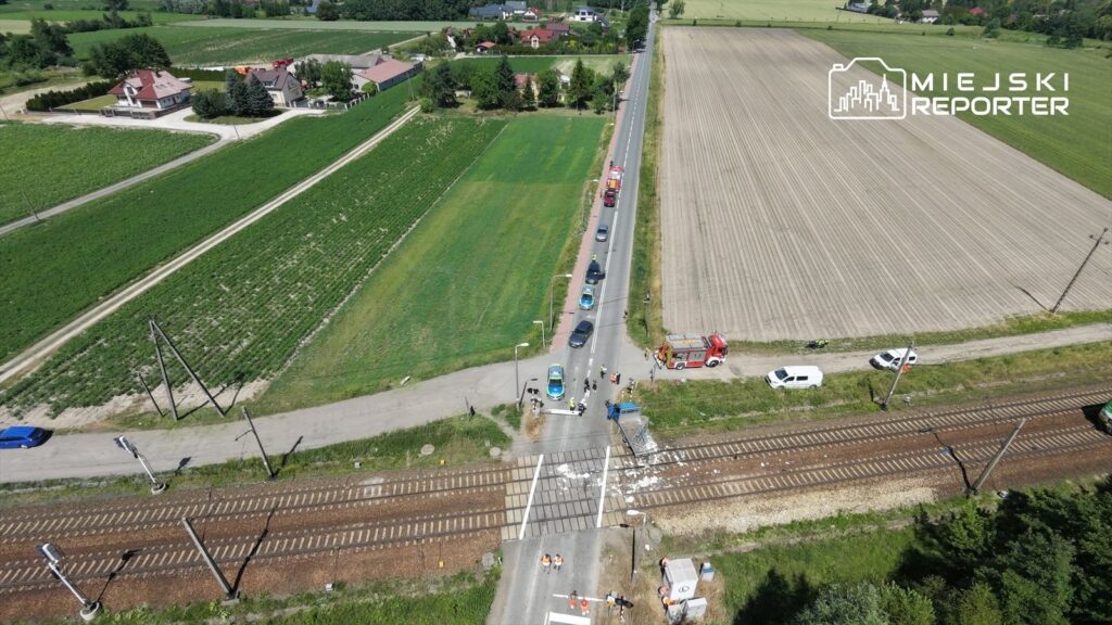 Pojazdy i służby ratunkowe zatrzymane przy przejeździe kolejowym na tle zielonych pól i zabudowań.