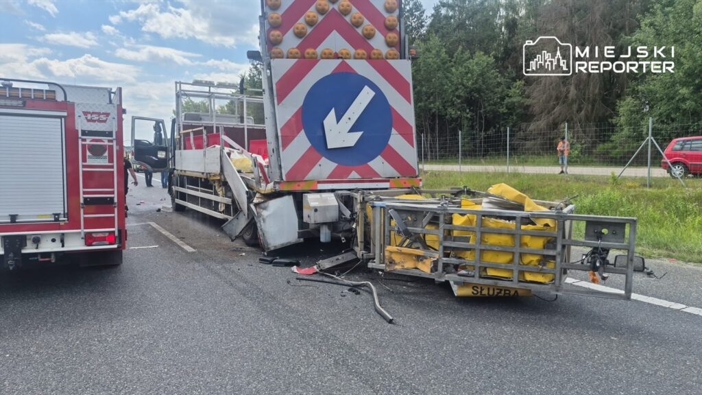 W miejscu wypadku znajduje się uszkodzony pojazd ciężarowy, obok którego pracują strażacy. Na drodze widoczne są pozostałości po kolizji.