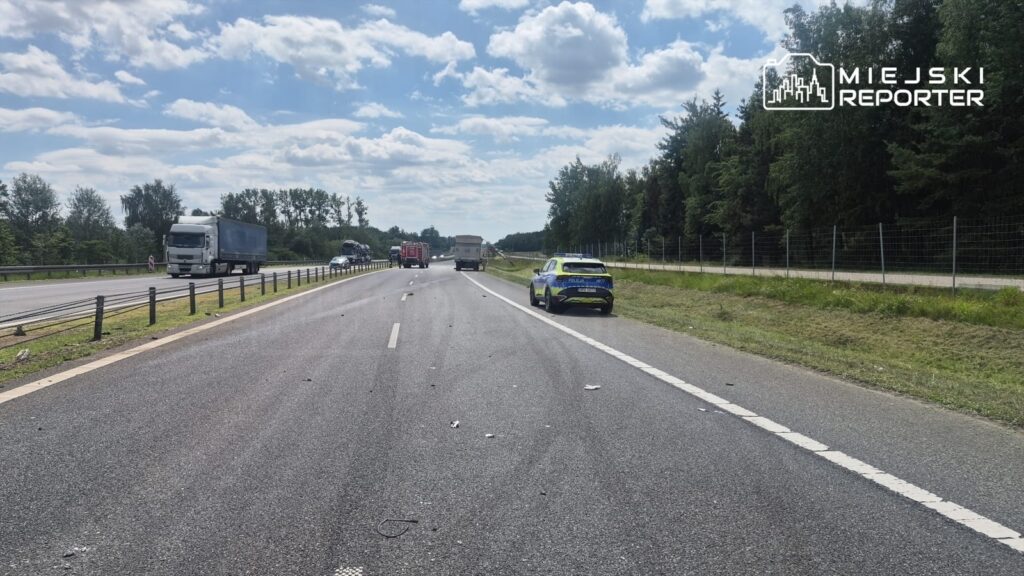 Na drodze widoczny wypadek, z uszkodzonym pojazdem na poboczu, obok stoi radiowóz policyjny.