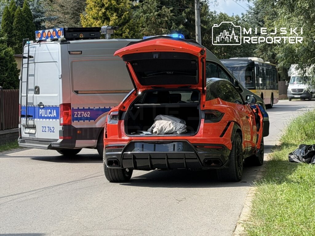 Czerwony samochód sportowy z otwartym bagażnikiem stoi na drodze obok policyjnego vana z włączonymi sygnałami świetlnymi.
