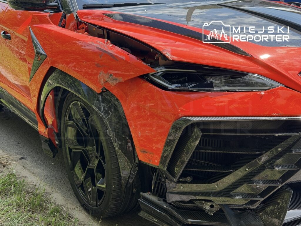 Uszkodzony czerwony samochód sportowy z zarysowaniami na nadwoziu, stojący na poboczu drogi otoczonej trawą.