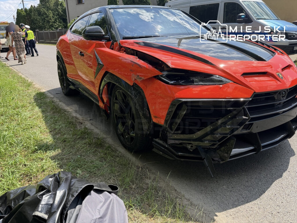 Czerwony SUV z uszkodzoną przednią częścią stoi na poboczu drogi, obok grupy osób oraz radiowozu policyjnego.