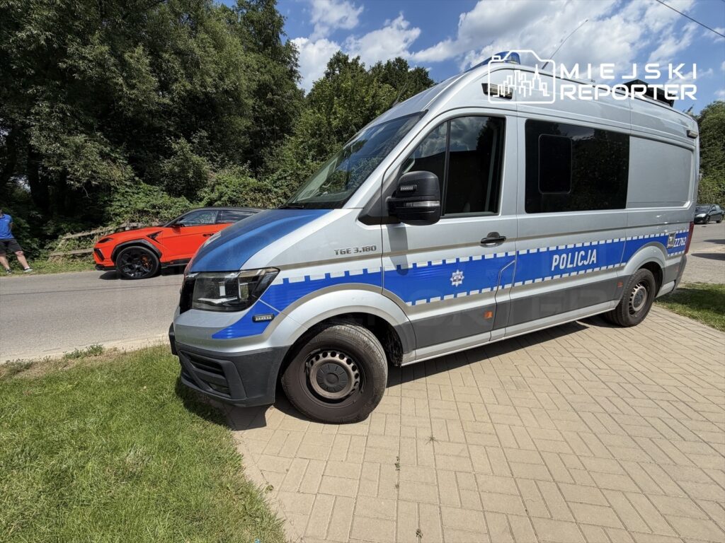 Nieoznakowany radiowóz policyjny zaparkowany na chodniku obok drogi, obok przejeżdżający czerwony samochód sportowy.