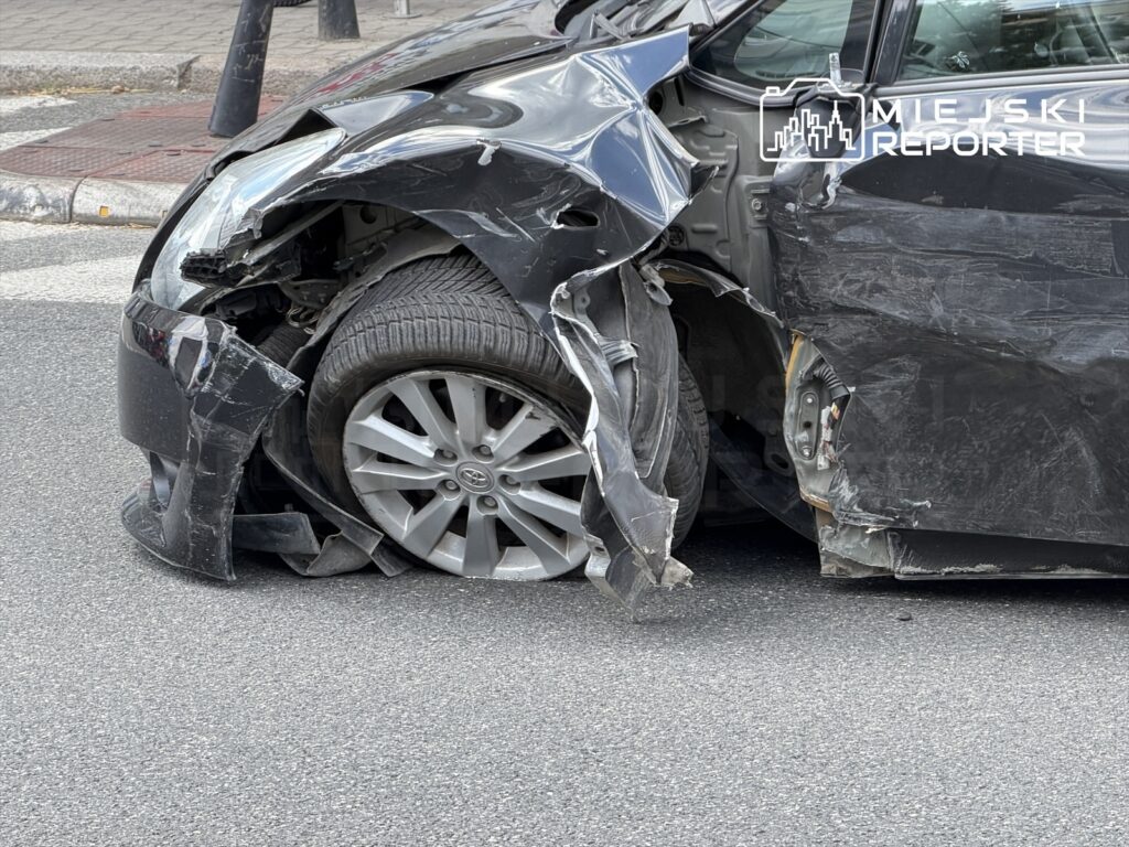 Uszkodzony czarny samochód osobowy z zniszczonym przodem i uszkodzoną oponą, zaparkowany na ulicy.