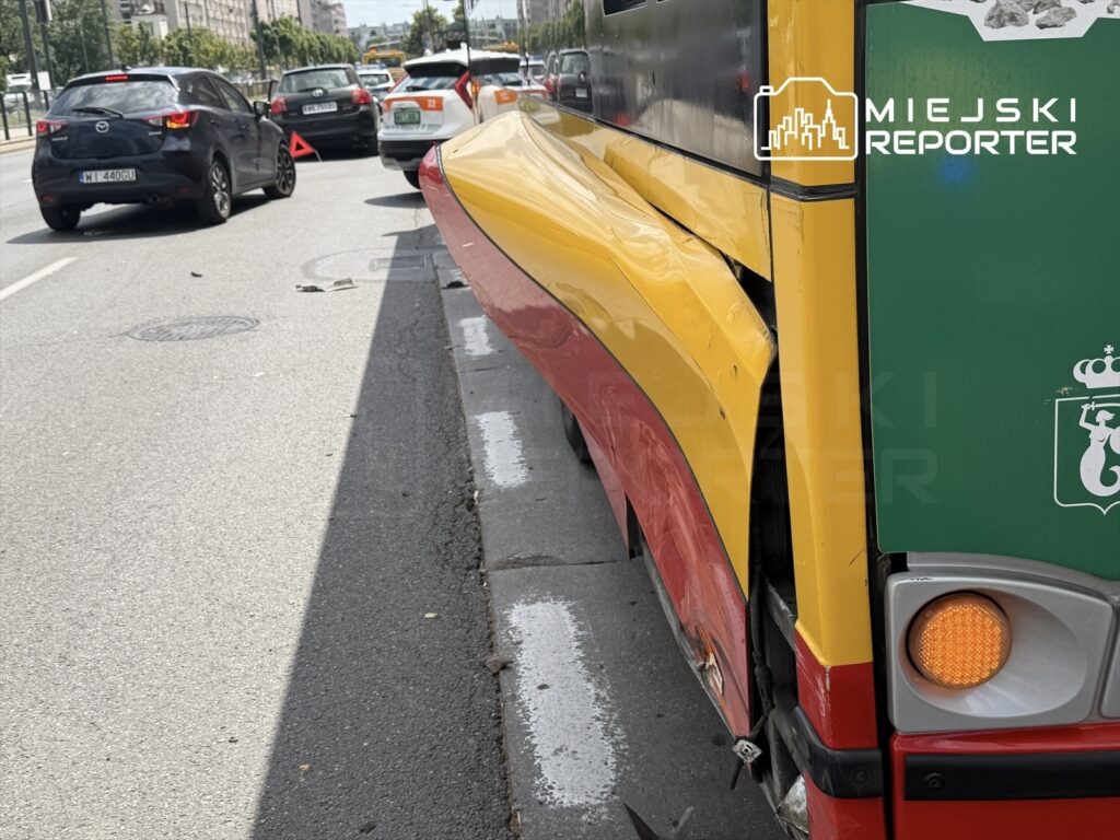 Zbliżenie na tył tramwaju z żółto-czerwonymi elementami, zaparkowanego obok ruchliwej ulicy z samochodami.