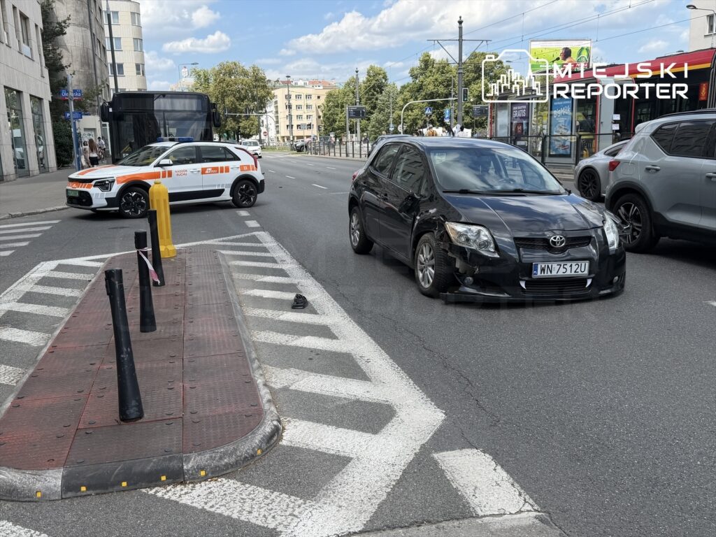 Czarny samochód osobowy z uszkodzeniem na przednim kole stoi na skrzyżowaniu, obok radiowozu z włączonymi sygnałami.