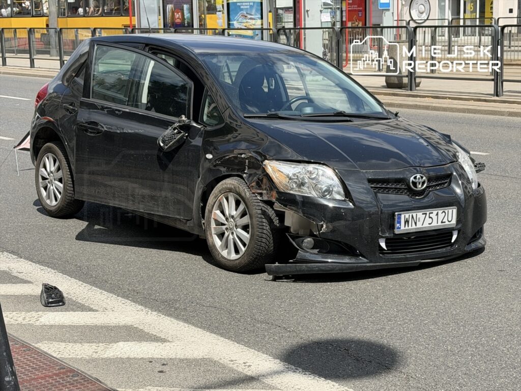 Czarny samochód osobowy z uszkodzeniami stoi na ulicy, obok leży fragment zderzaka.