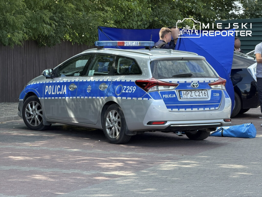 Policyjny radiowóz zaparkowany na ulicy, obok stoiska zabezpieczonego niebieskim namiotem.